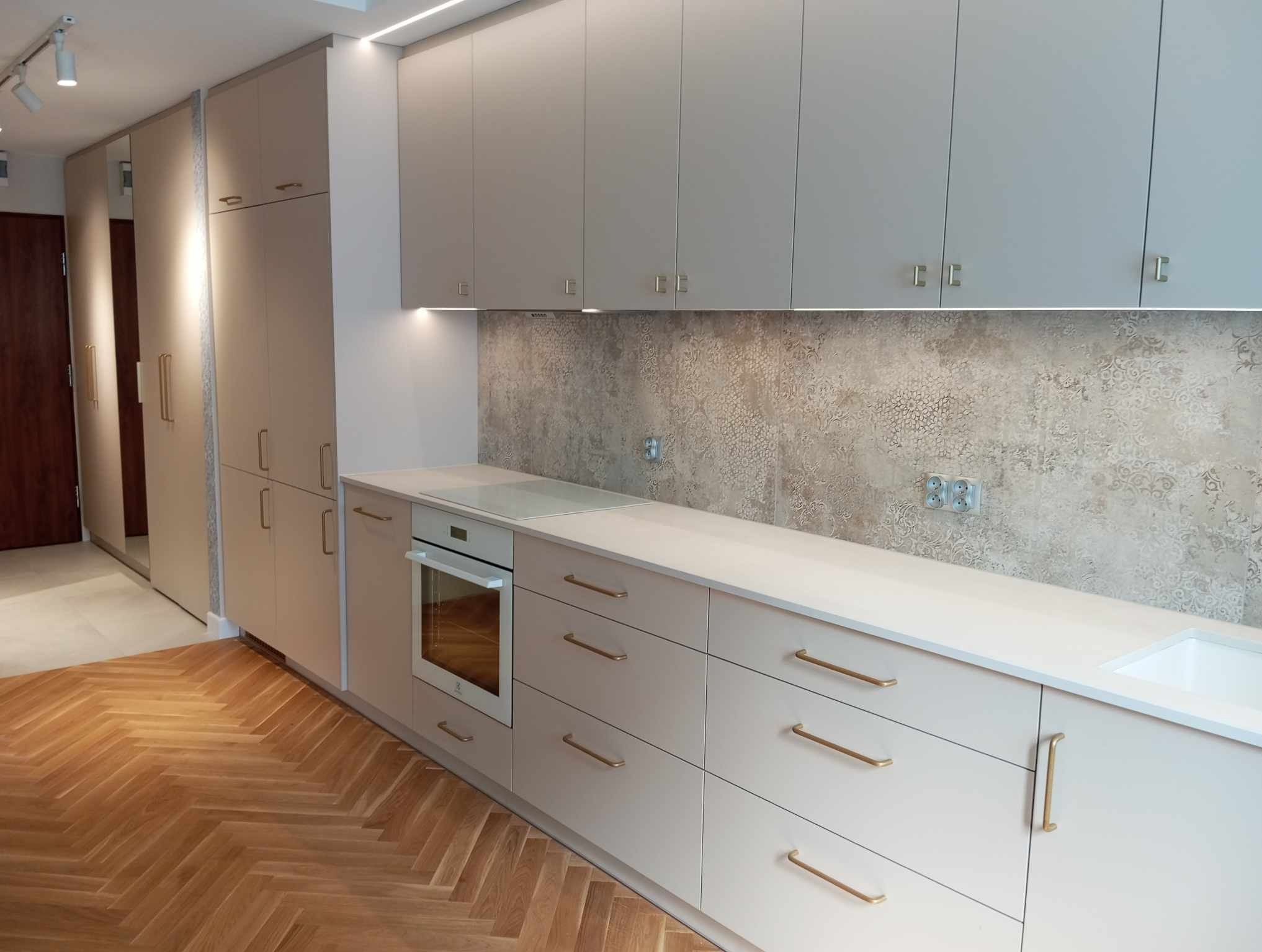 Jasna kuchnia z drewnianą podłogą w jodełkę, beżowe fronty szafek z poziomymi uchwytami, piekarnik w zabudowie, dekoracyjna płytka nad blatem, oświetlenie LED podszafkowe.