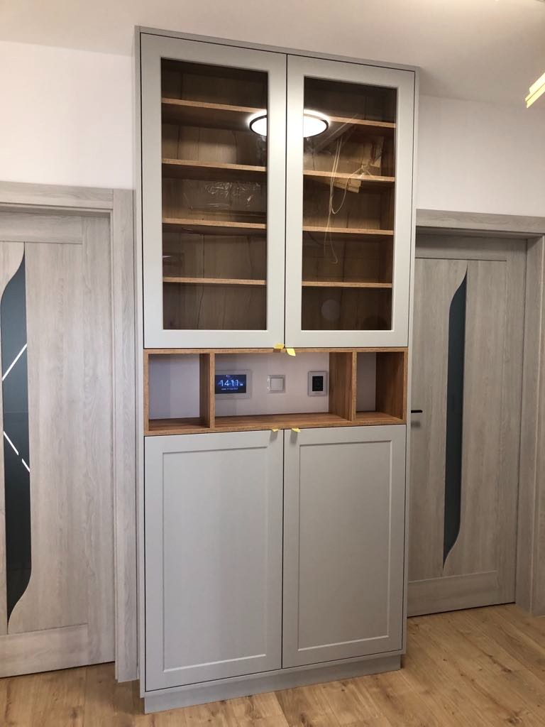 Wysoka, szara szafa z przeszklonymi górnymi drzwiczkami i drewnianymi półkami wewnątrz, zintegrowana z panelem sterowania domem i oświetleniem LED, umieszczona między dwiema szarymi drzwiami.