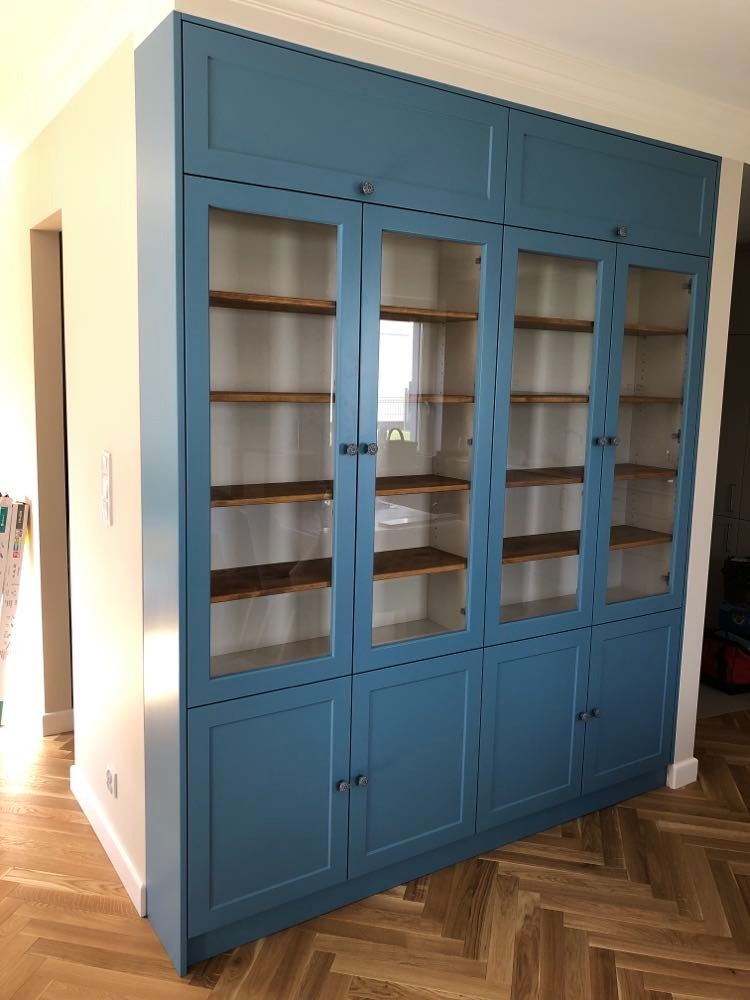 Niebieska zabudowa meblowa z przeszklonymi witrynami i drewnianymi półkami, wkomponowana we wnękę ściany, na podłodze parkiet w jodełkę.