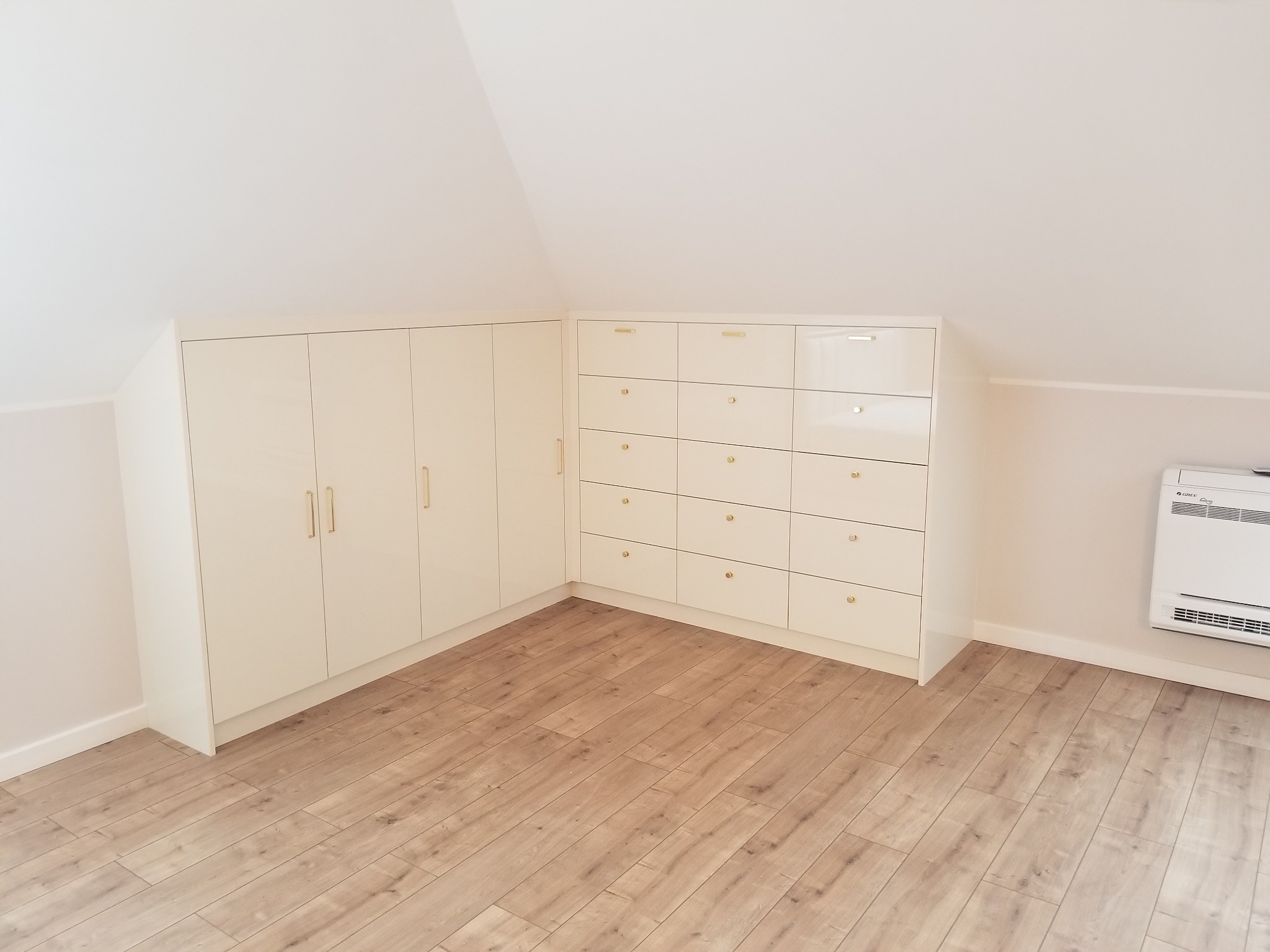 Białe szafy i komody z błyszczącymi frontami i złotymi uchwytami, zamontowane pod skosem dachu na poddaszu, z drewnianą podłogą.