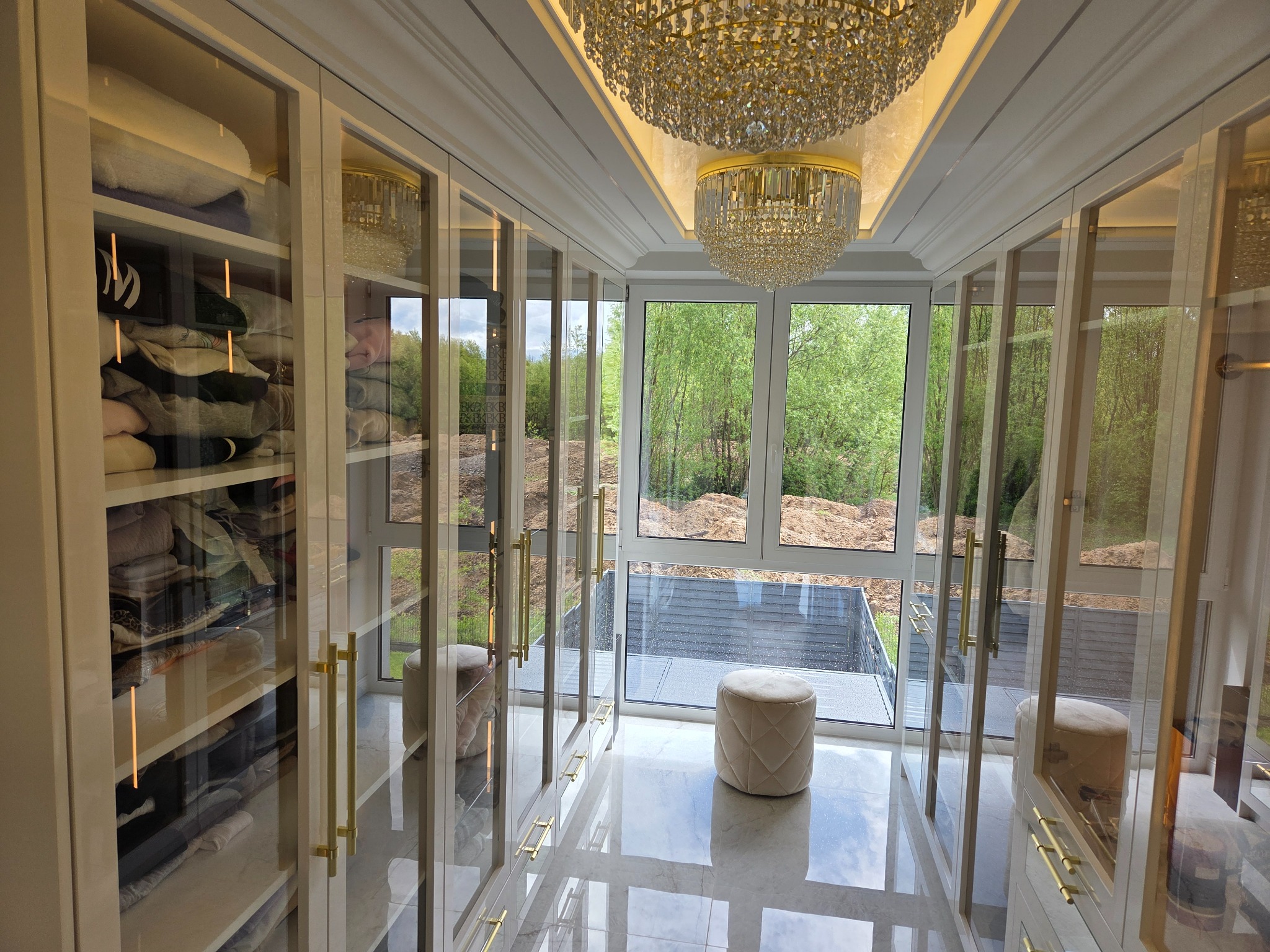 Elegancka garderoba z przeszklonymi szafami, złotymi uchwytami i kryształowym żyrandolem. Widok na zieleń za oknem.