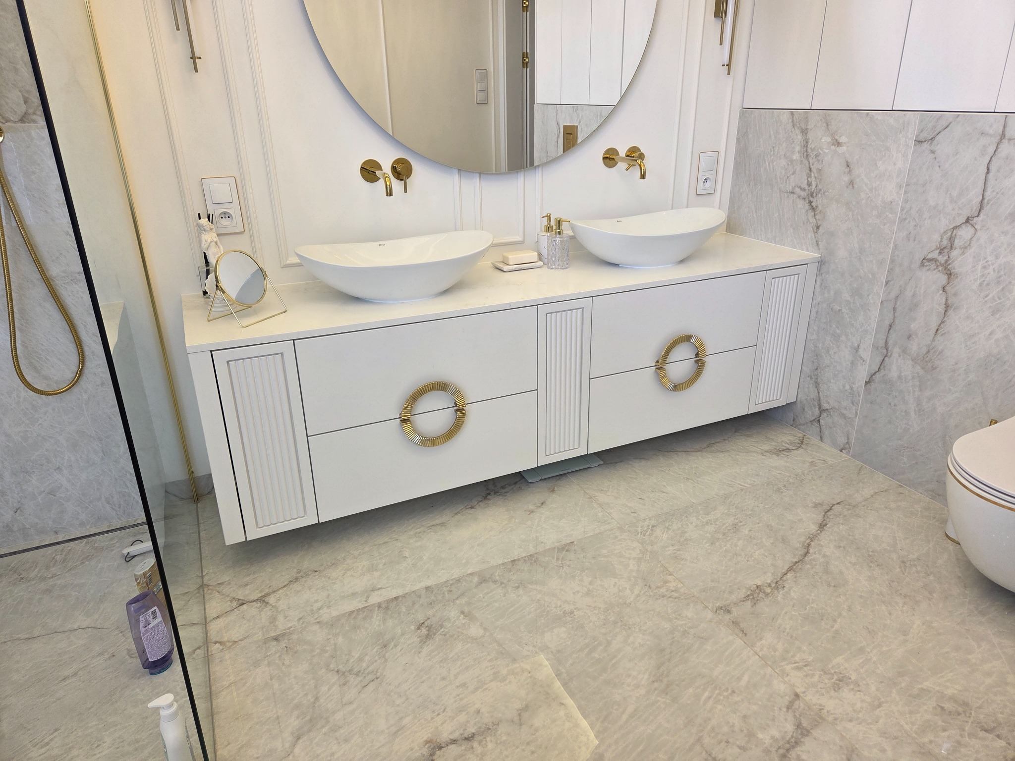 Elegancka łazienka z podwójną umywalką na białej szafce z ozdobnymi, złotymi uchwytami. Okrągłe lustro i marmurowe płytki dodają luksusowego charakteru.