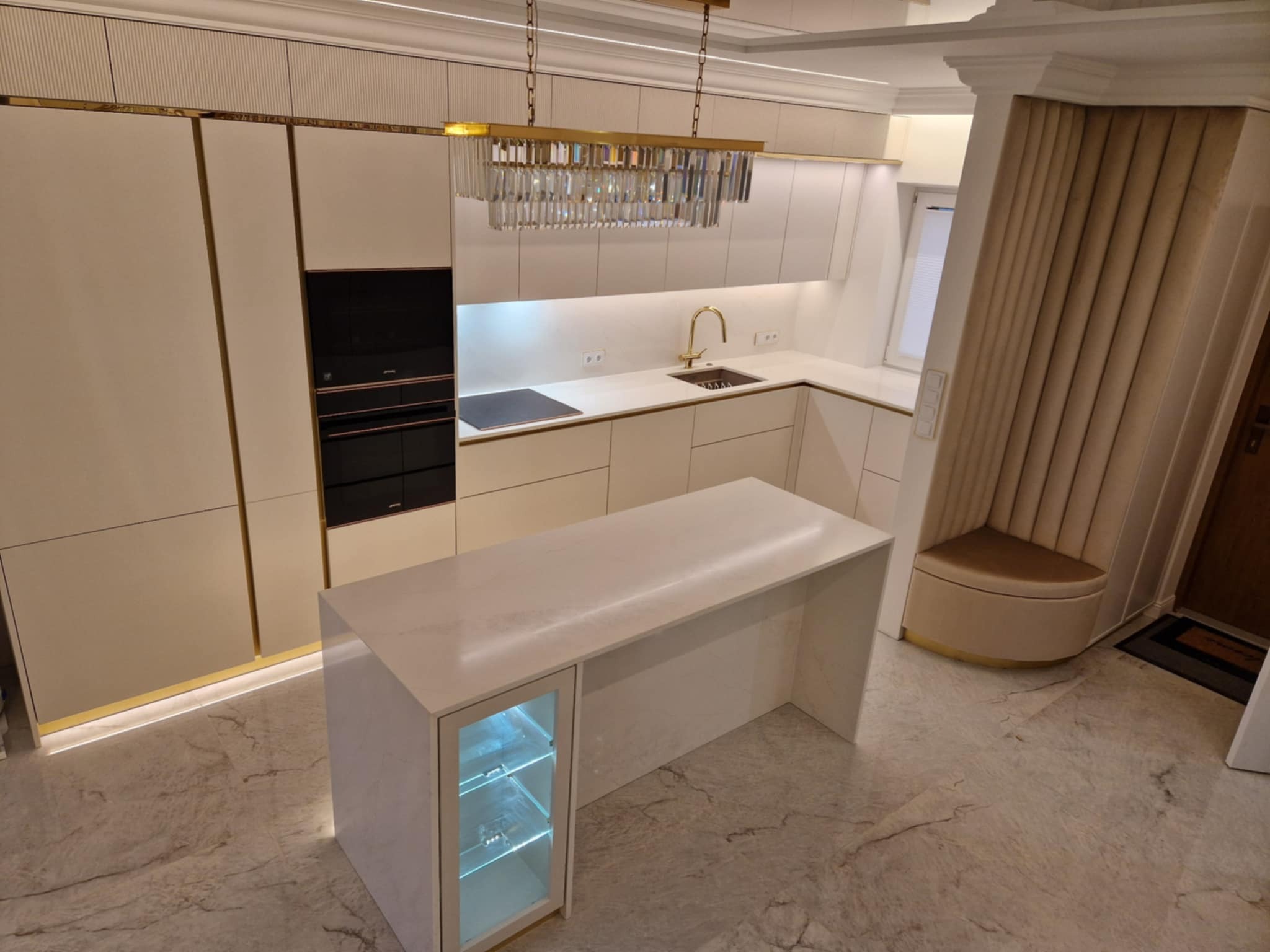 Elegancka kuchnia w odcieniach beżu z wyspą i oświetleniem kryształowym. Fronty szafek zdobione złotymi detalami, marmurowa podłoga. Wbudowany sprzęt AGD.