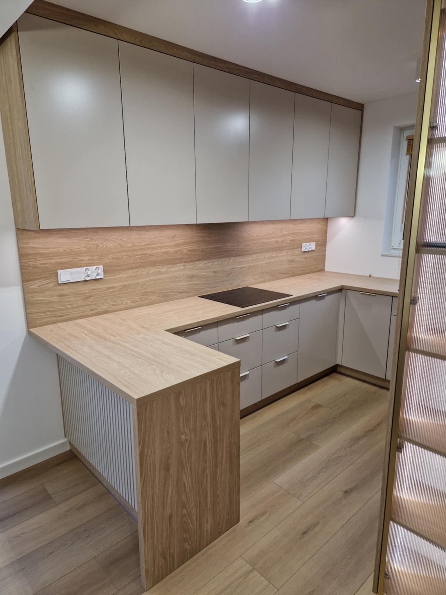 Jasna kuchnia z drewnianym blatem i frontami szafek w odcieniach beżu i szarości. Widoczna płyta indukcyjna i ryflowany panel boczny. Całość w nowoczesnym stylu.