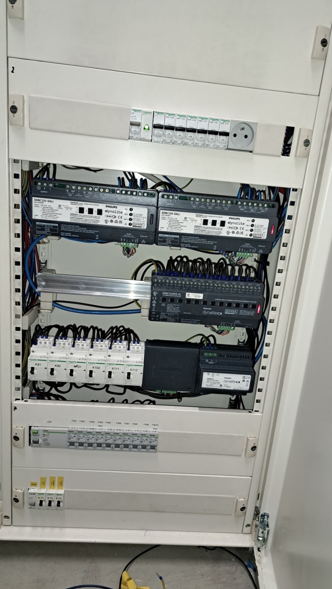 Otwarta szafa elektryczna z zamontowanymi modułami Philips Dynalite, bezpiecznikami i okablowaniem, widoczne elementy sterujące oświetleniem DALI i moduły przekaźnikowe.