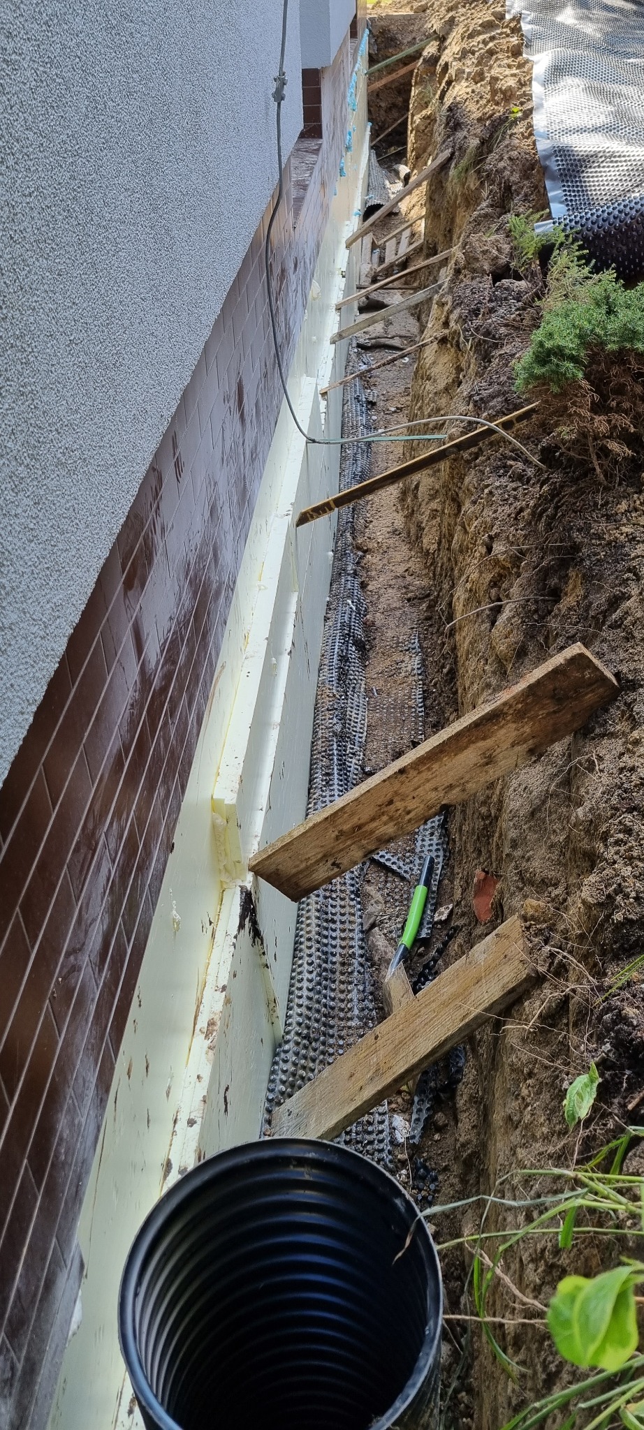 Fundament ocieplony styropianem i zabezpieczony folią kubełkową w wykopie. Widoczne drewniane podpory i fragment rury drenażowej.