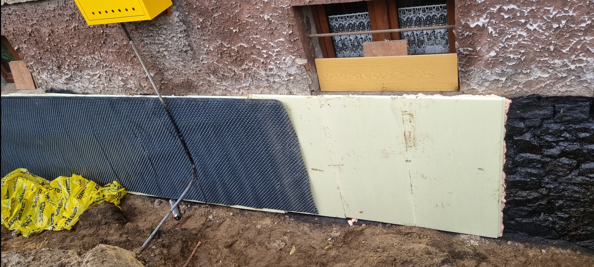 Ocieplenie fundamentów budynku w Będzinie: styropian, folia kubełkowa i izolacja bitumiczna. Widoczne żółte worki z materiałem izolacyjnym.