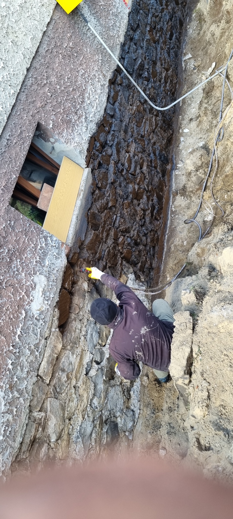 Natrysk hydroizolacji na nierówną ścianę fundamentu przez pracownika w rękawicach, widok z góry. Częściowo zaizolowana ściana, przewody i wykop.