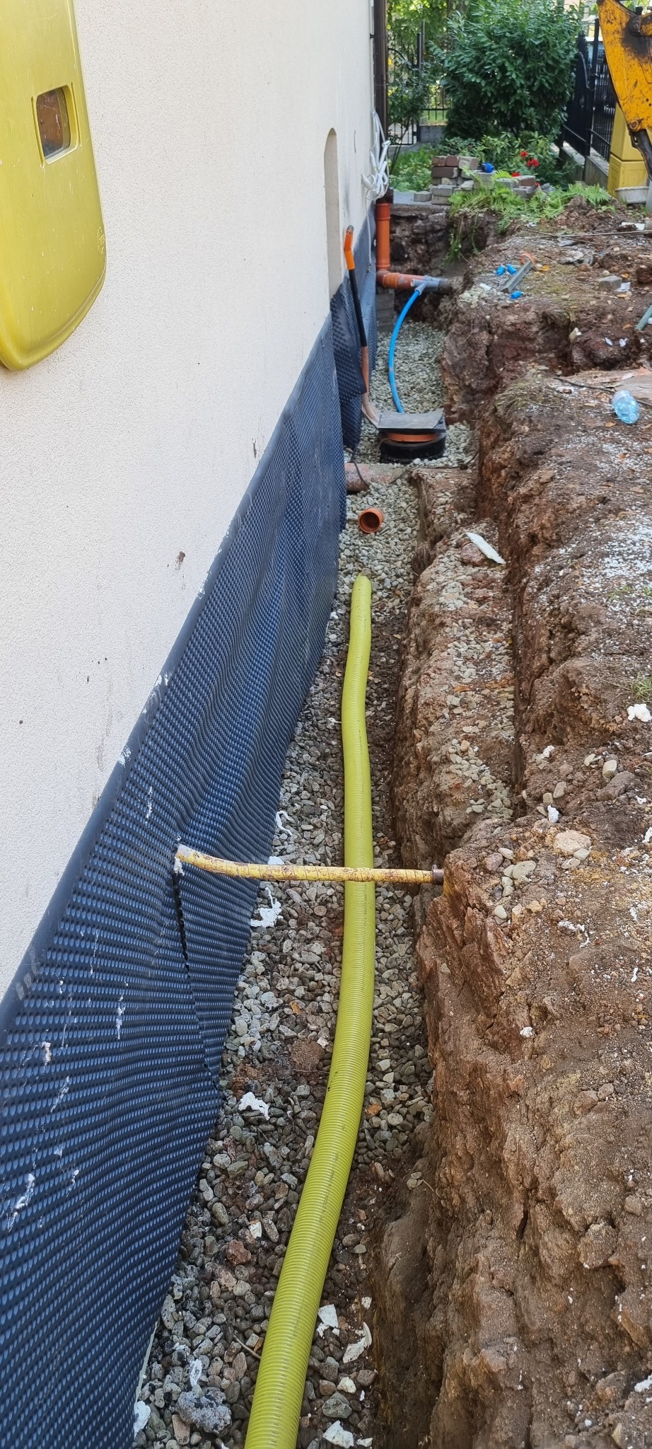 Fundament zabezpieczony folią kubełkową i drenażem w wykopie. Widoczne rury odprowadzające wodę. Prace ziemne w toku, fragment koparki w tle.