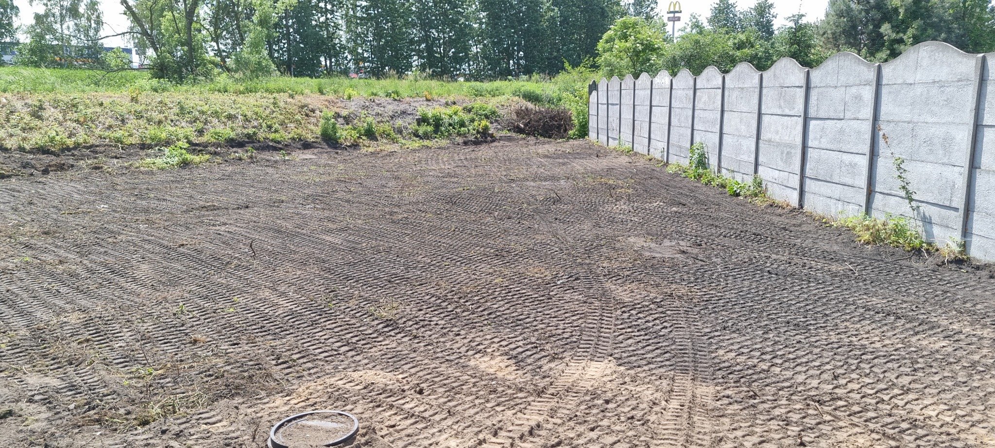 Przygotowany teren pod budowę z widocznymi śladami opon, wyrównana ziemia, betonowe ogrodzenie w tle i częściowo zachowana roślinność na obrzeżach.