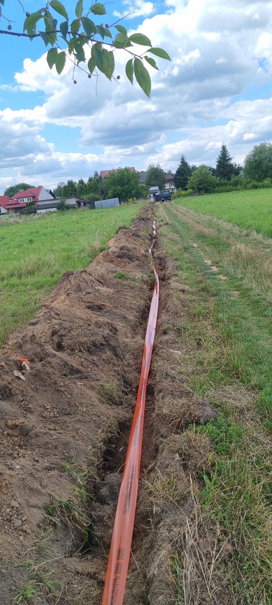 Długi, wąski wykop w trawiastym terenie, zabezpieczony pomarańczową taśmą ostrzegawczą, prowadzący w kierunku zabudowań na horyzoncie pod błękitnym, obłocznym niebem.