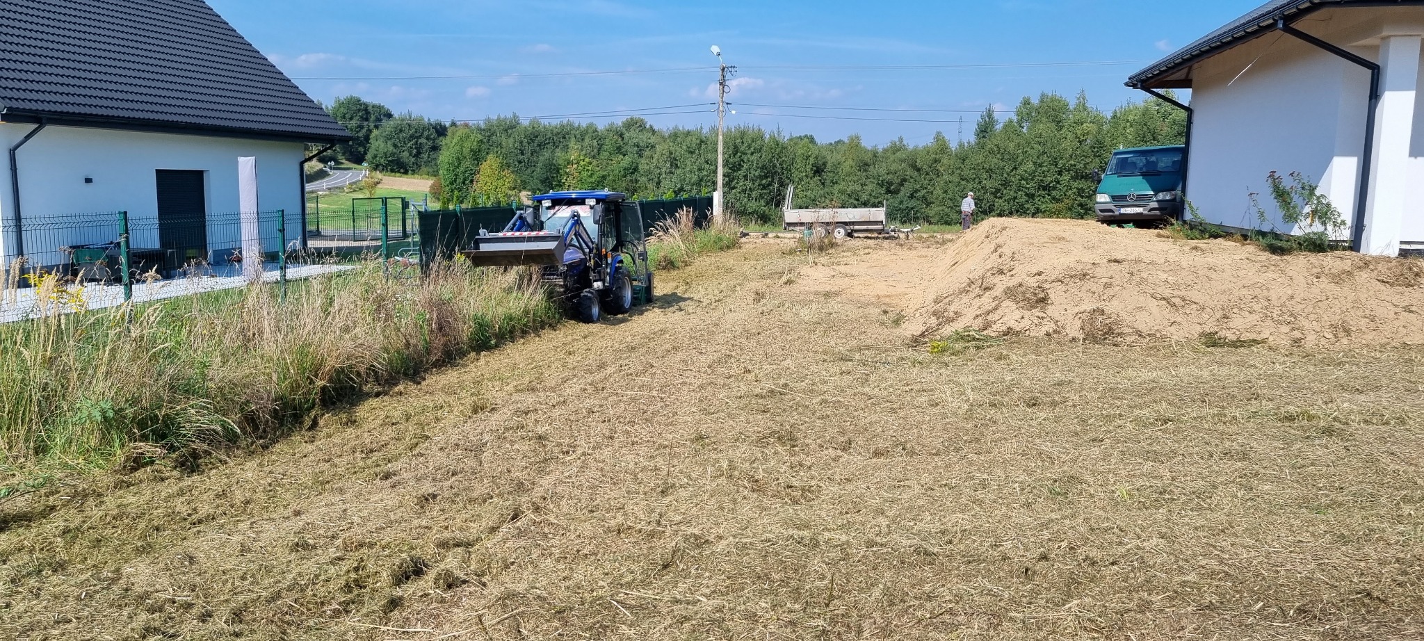 Niebieski traktor z łyżką wyrównuje teren działki, w tle widoczny dom z ciemnym dachem, sterta ziemi i częściowo zasłonięty bus dostawczy.