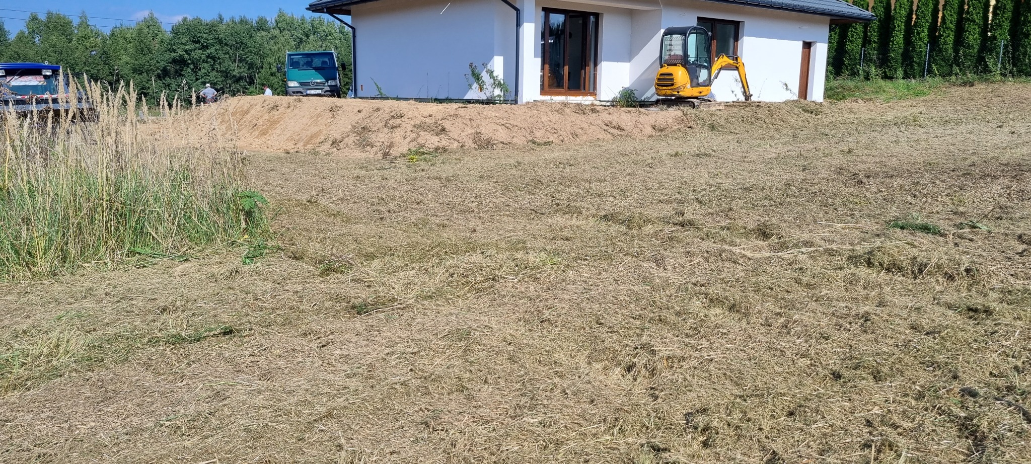 Żółta mini koparka stoi na trawniku przed białym domem, za nią usypana pryzma piasku, w tle widoczne dwa samochody dostawcze i drzewa.