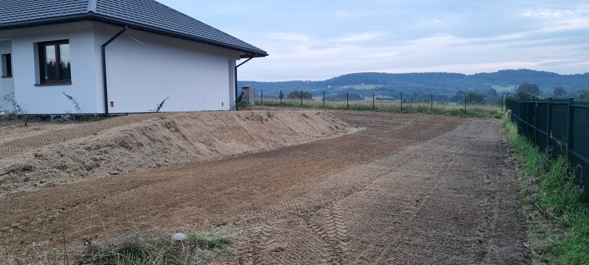 Przygotowany teren pod budowę domu jednorodzinnego, świeżo wyrównana ziemia z widocznymi śladami opon, hałda piasku przy ścianie budynku, w tle zalesione wzgórza.