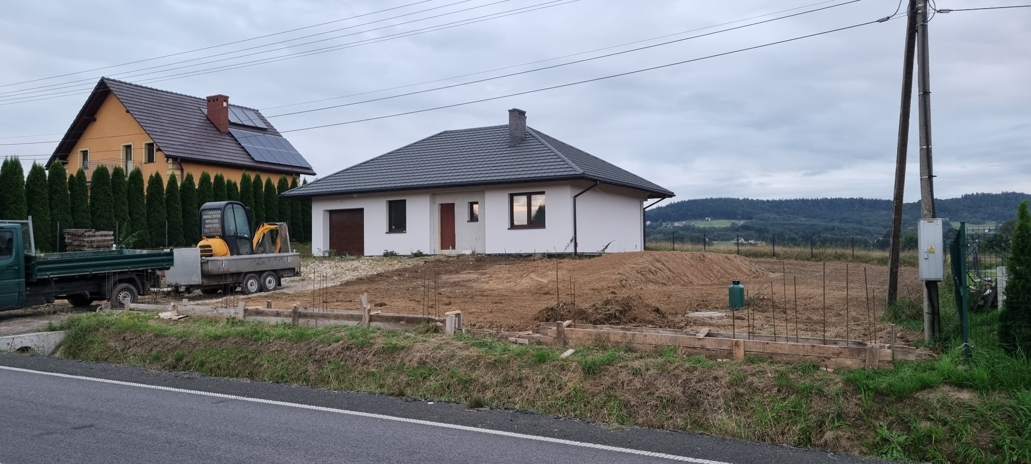 Nowo wybudowany, parterowy dom z szarą dachówką, widoczny z perspektywy ulicy, z przygotowanym terenem pod fundamenty, drewnianym szalunkiem i stertą ziemi. Na lawecie stoi mała koparka.