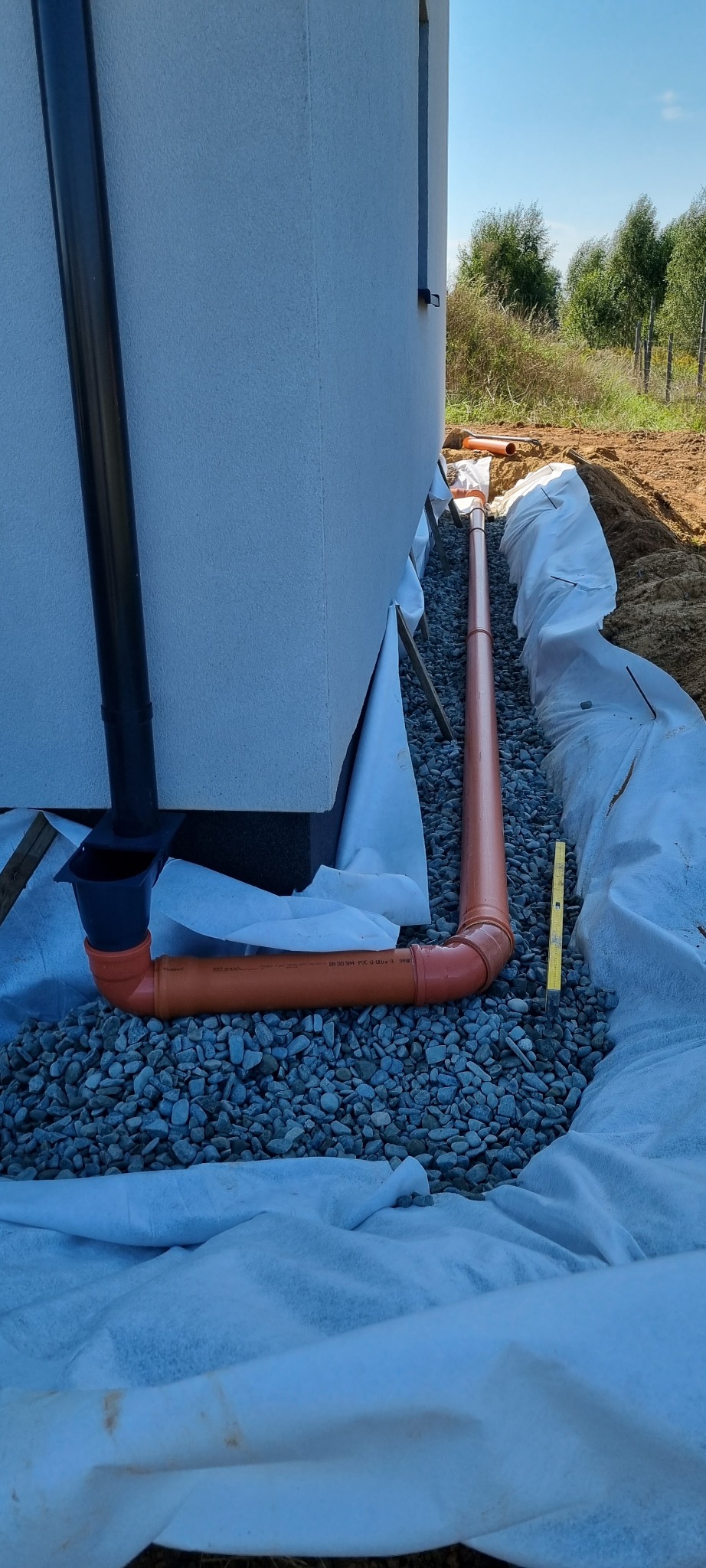 Pomarańczowa rura drenażowa ułożona w wykopie wyłożonym białą geowłókniną, otoczona szarym żwirem, widoczny fragment budynku i pionowa rura spustowa