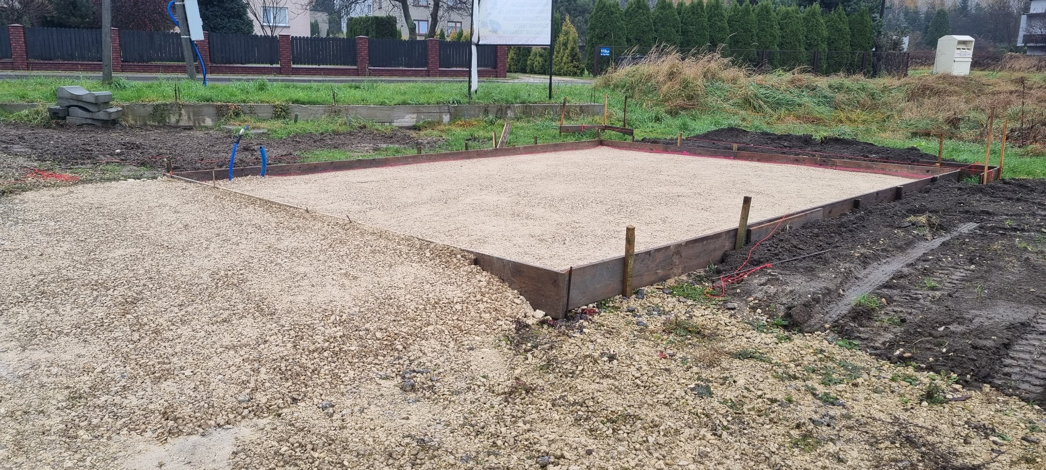 Przygotowany teren pod wylewkę fundamentów z drewnianym szalunkiem i podsypką żwirową, widoczne ślady opon i elementy instalacji.