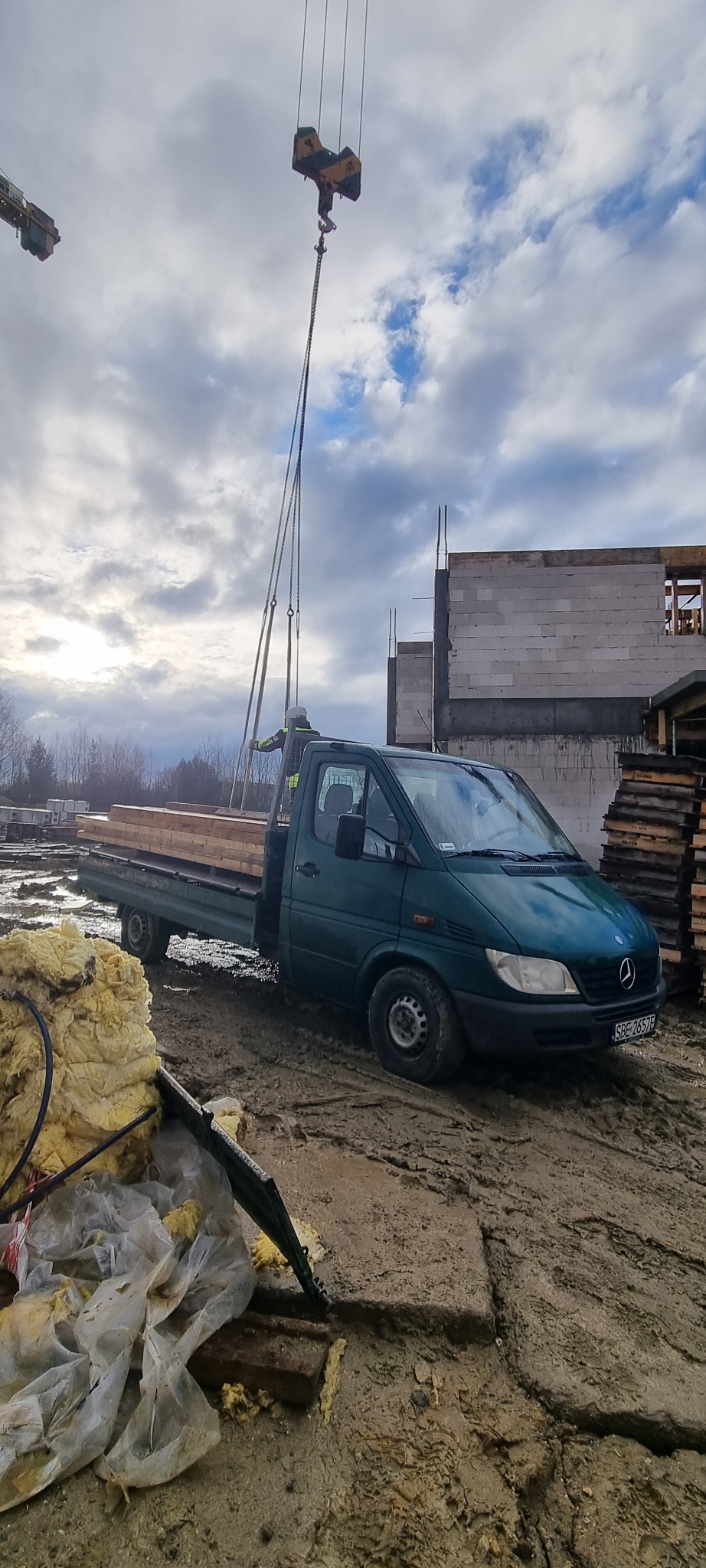 Zielony samochód dostawczy z deskami na pace, obok budowa w trakcie realizacji, w tle dźwig podnosi materiały budowlane, błotniste podłoże.