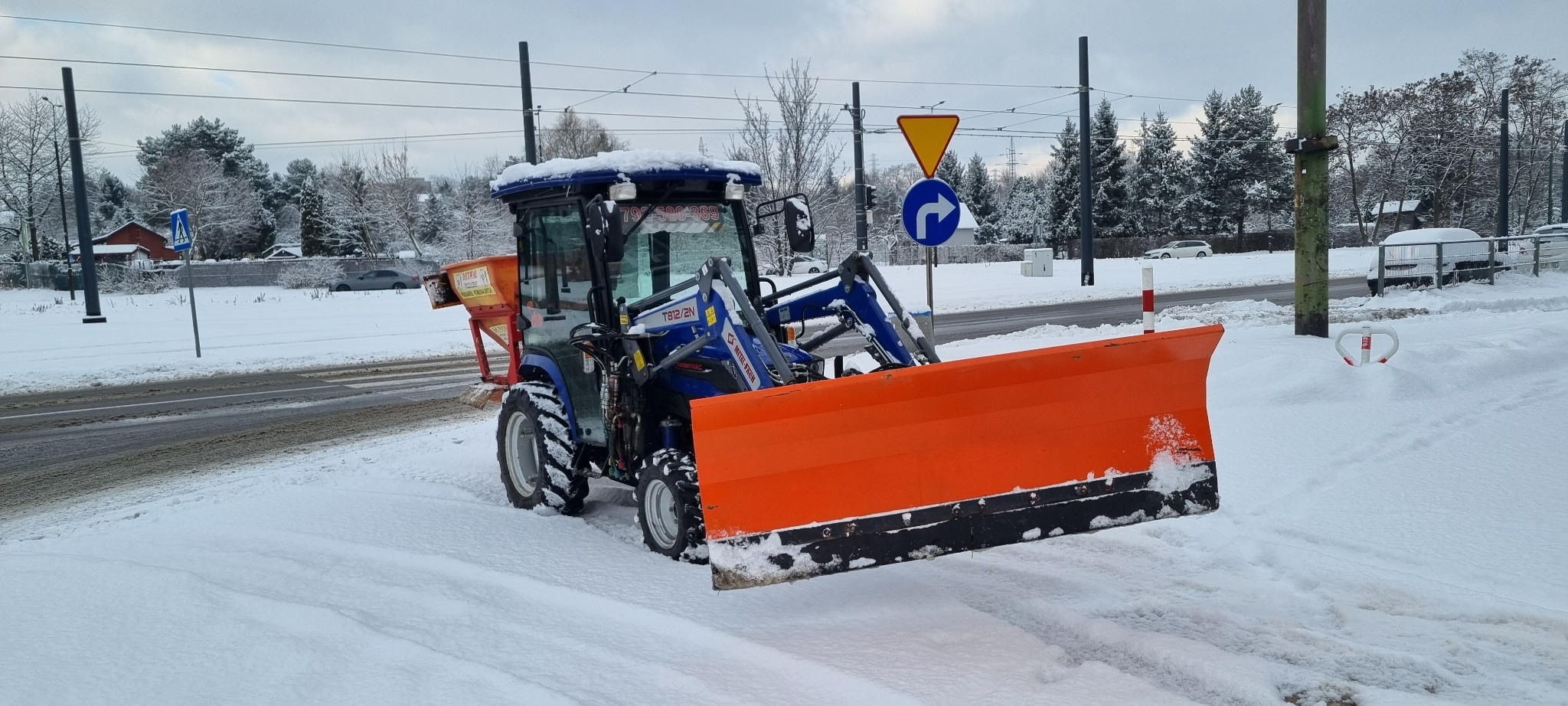 Niebieski traktor z pomarańczowym pługiem do odśnieżania, zaparkowany na zasypanym śniegiem poboczu drogi, z posypywarką piasku na tyle, w tle widoczne drzewa i znaki drogowe.