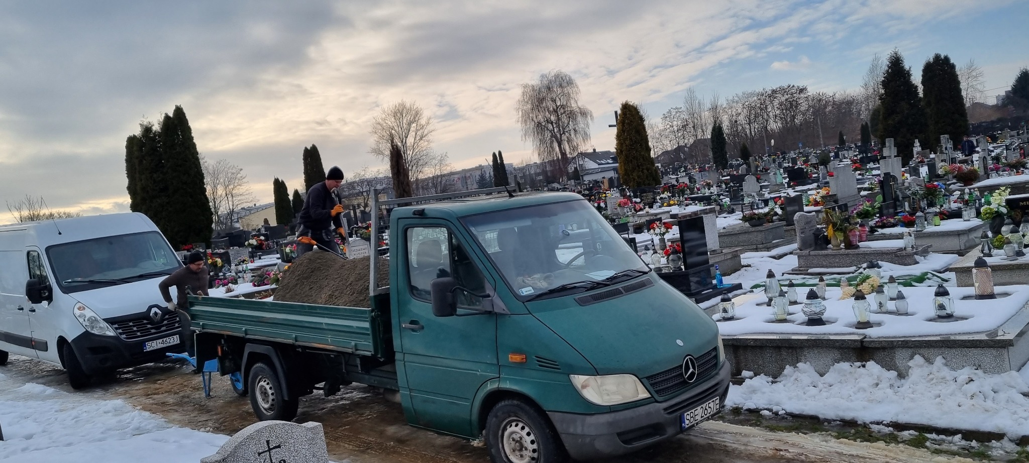 Pracownik łopatą wyrównuje ziemię w skrzyni zielonego samochodu ciężarowego Mercedes, na tle zimowego krajobrazu cmentarza z nagrobkami pokrytymi śniegiem.