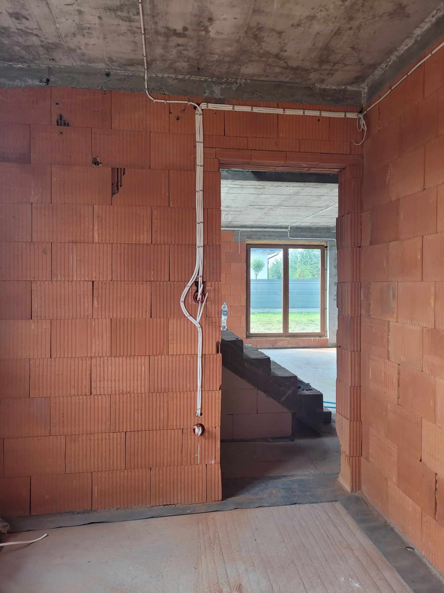 Surowy stan wnętrza budynku w trakcie budowy, widoczne przewody elektryczne prowadzone po ścianach z czerwonej cegły, surowy betonowy strop, okno z widokiem na ogród.
