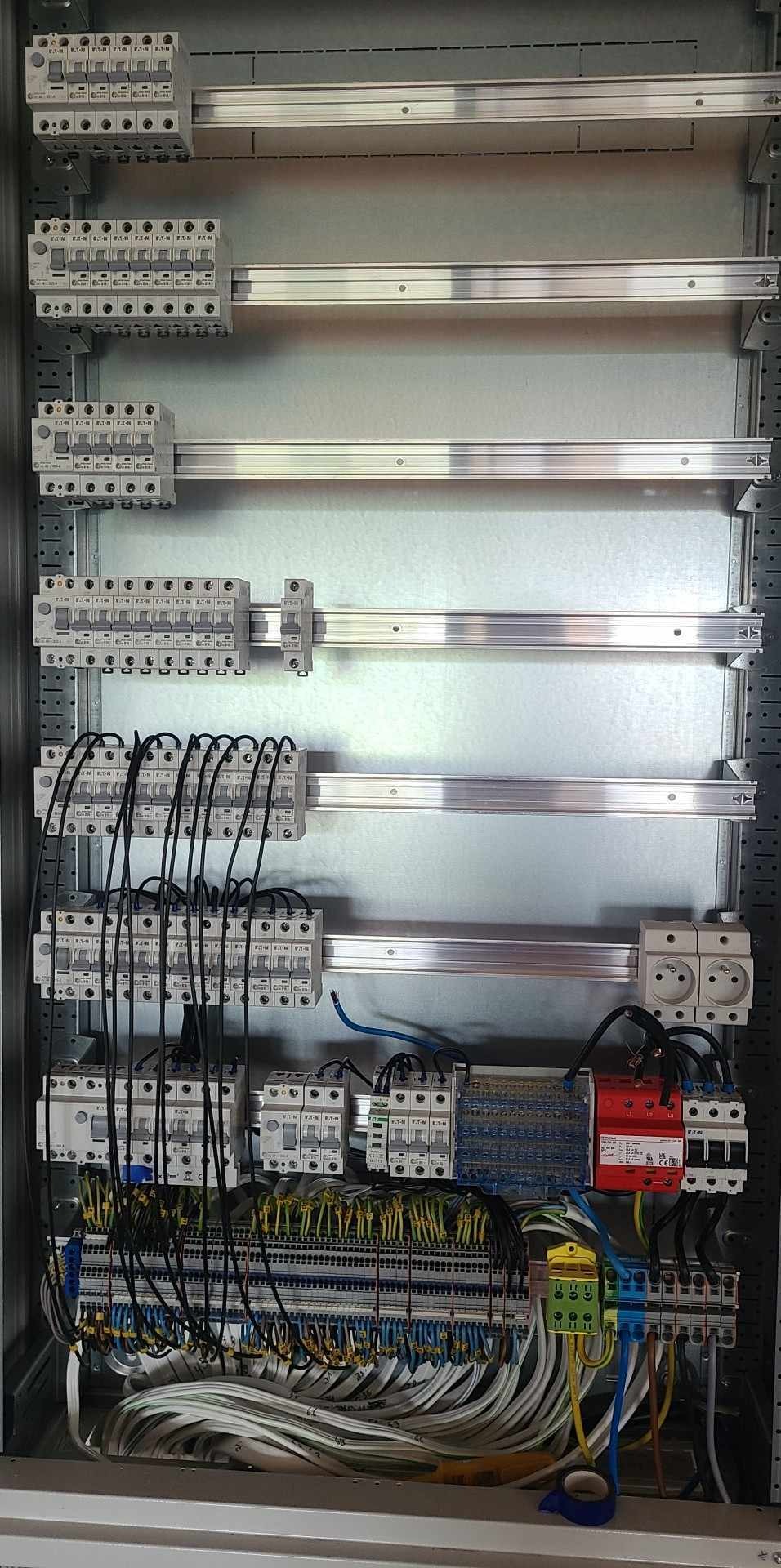 Rozdzielnica elektryczna z zamontowanymi bezpiecznikami, listwami zaciskowymi i przewodami o różnych kolorach, widoczne gniazdka elektryczne.