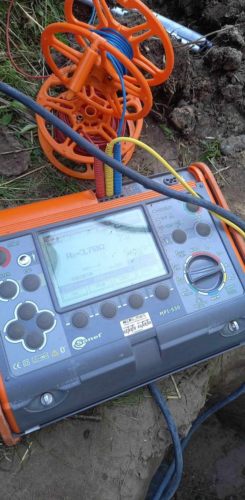 Miernik Sonel MPI-530 na ziemi z widocznym wynikiem pomiaru rezystancji uziemienia Re=3,700 Ω, obok rozwinięty bęben z przewodami pomiarowymi w kolorach niebieskim, żółtym i czerwonym.