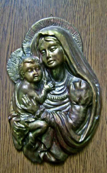 Malowany odlew gipsowy przedstawiający Matkę Boską z Dzieciątkiem, patynowany na złoto i srebro, zawieszony na drewnianej ścianie.