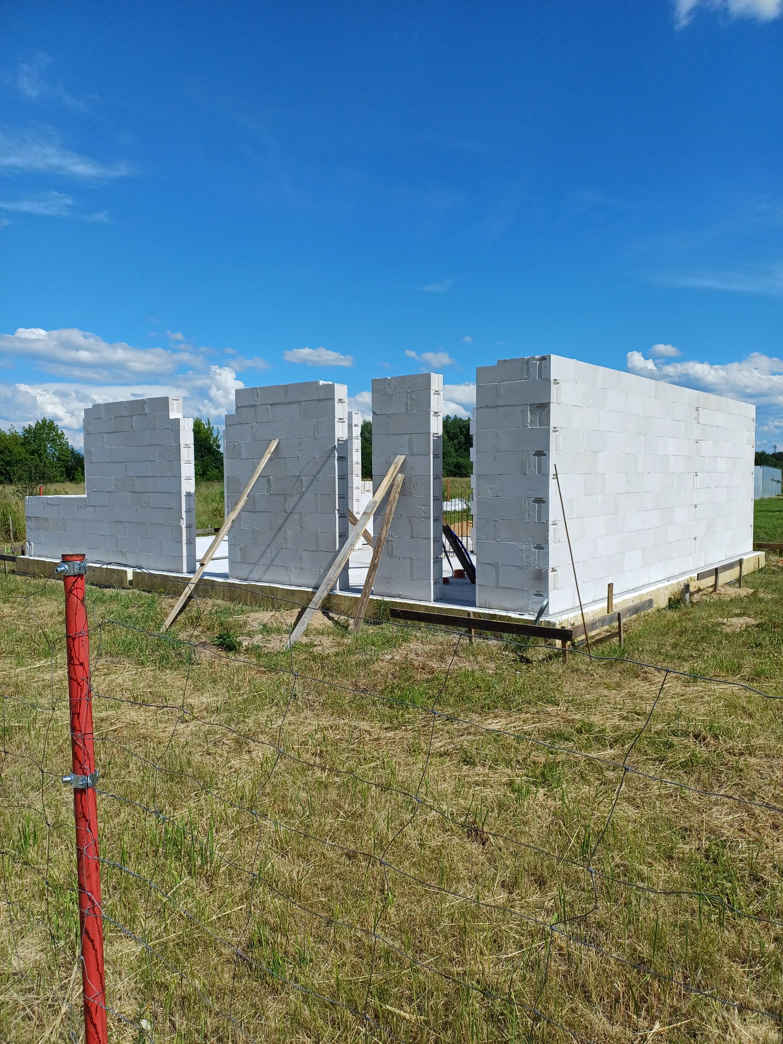 Wznoszone ściany domu z białych bloczków na fundamencie, podparte drewnianymi stemplami, widok przez siatkę ogrodzeniową na tle błękitnego nieba.