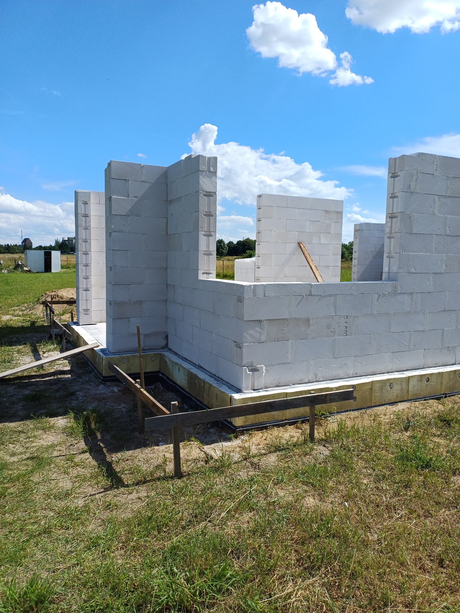 Wznoszone ściany domu z białych bloczków betonowych na fundamencie z izolacją termiczną, widoczne drewniane podpory i elementy szalunku, w tle krajobraz polny pod błękitnym, lekko zachmurzonym niebem.