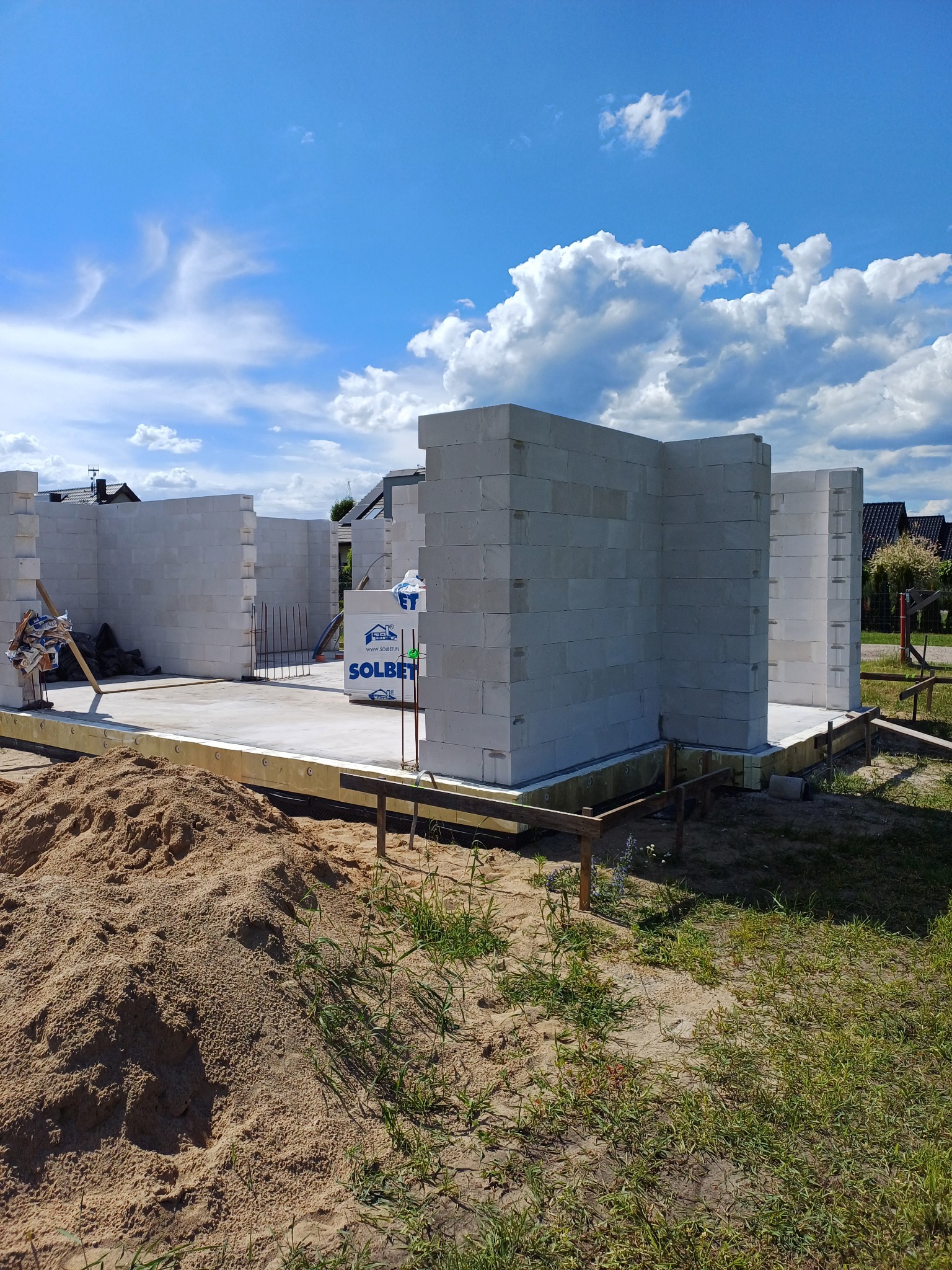 Wznoszenie ścian parterowego domu z betonu komórkowego Solbet, widoczne palety z bloczkami, fundamenty i drewniana konstrukcja szalunkowa, błękitne niebo z obłokami w tle.