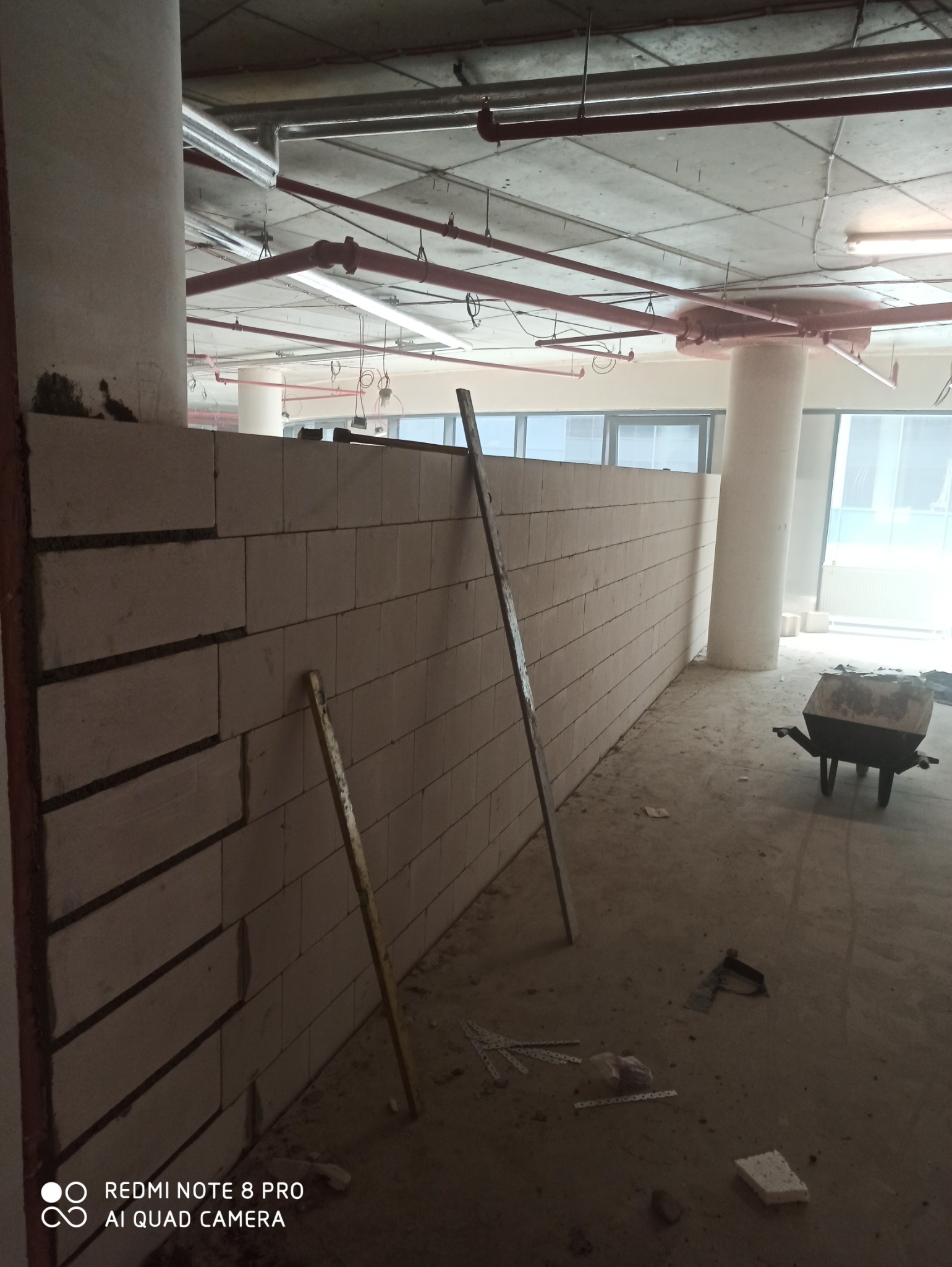 Wznoszona ściana z bloczków betonu komórkowego na surowym betonowym podłożu wewnątrz budynku, podparta dwiema łatami, z widoczną taczką w tle i instalacją pod sufitem.