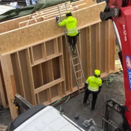 Montaż ściany szkieletowej domu modułowego z użyciem dźwigu, pracownicy w odblaskowych ubraniach na drabinie, widoczne elementy konstrukcyjne.