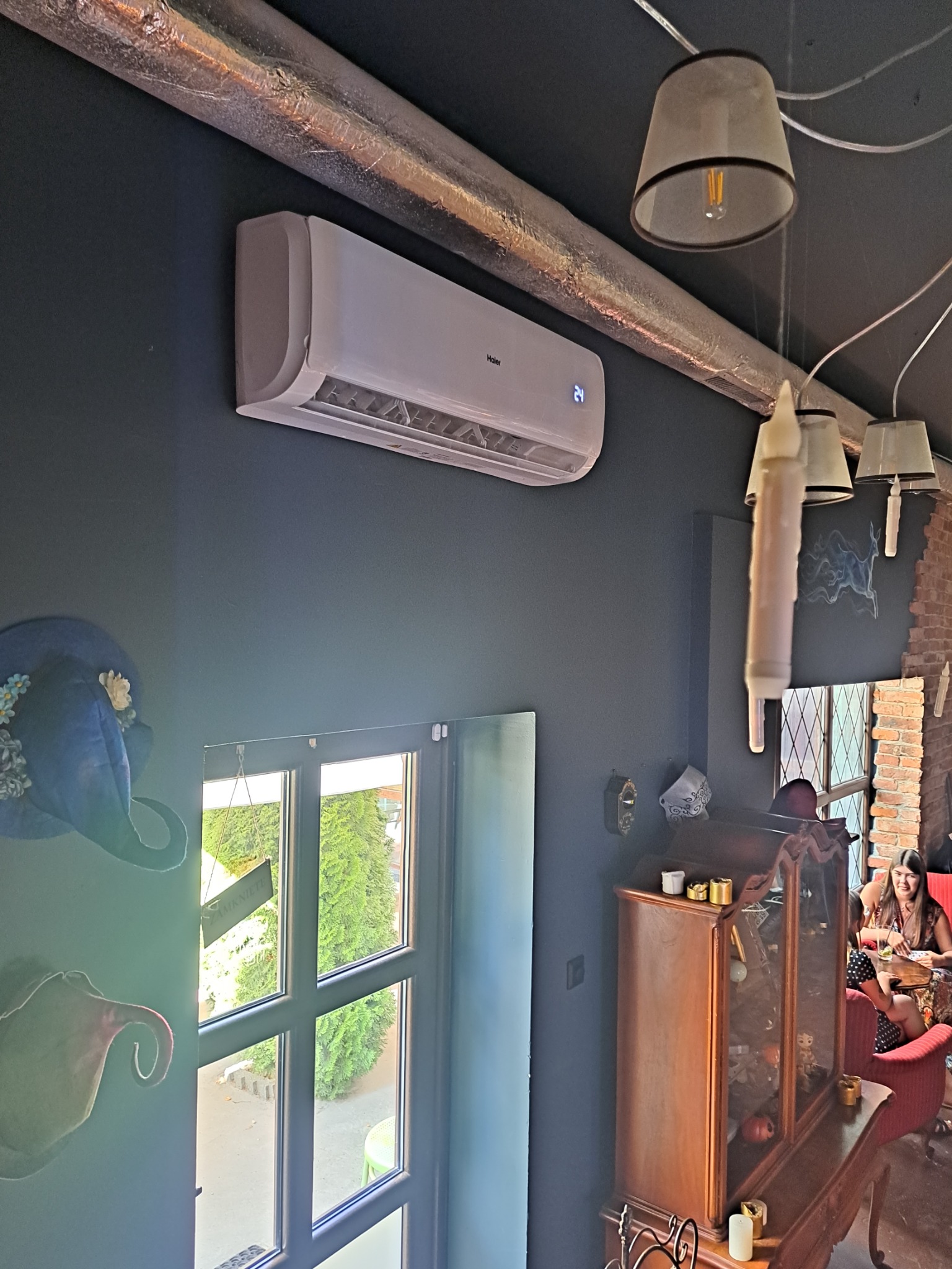 Biała klimatyzacja Haier na ciemnej ścianie lokalu, widoczna rura wentylacyjna, lampy wiszące, okno z widokiem na zewnątrz i fragment wnętrza z klientami.