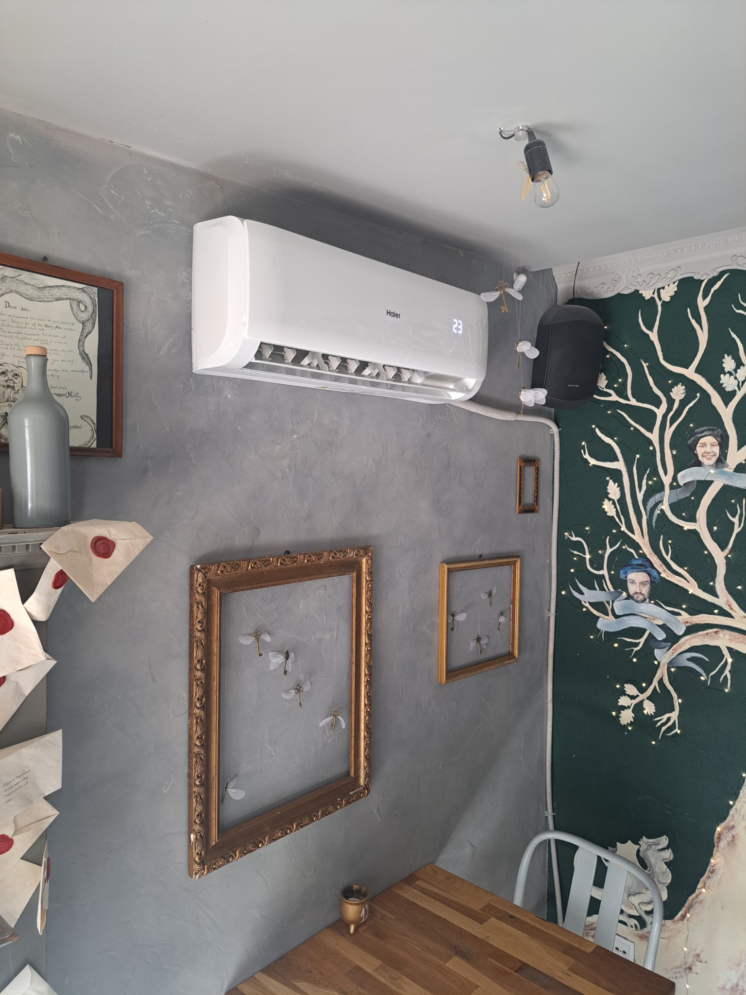 Biały klimatyzator Haier zamontowany na szarej ścianie w pomieszczeniu o wystroju inspirowanym stylem magicznym, widoczne dekoracje nawiązujące do tego stylu, takie jak obrazy ze skrzydlatymi...