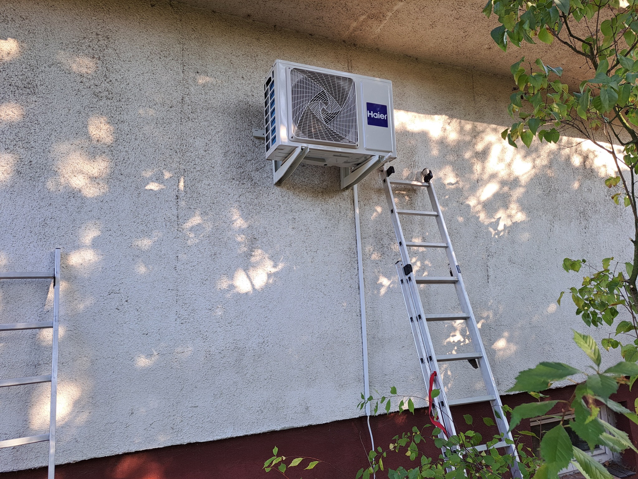 Zewnętrzna jednostka klimatyzacji Haier zamontowana na ścianie budynku, z drabiny oparta o ścianę i widoczne cienie drzew.
