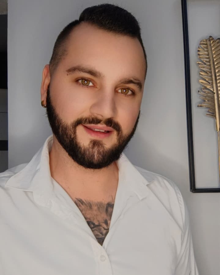 Portret uśmiechniętego mężczyzny z brodą i tatuażem na klatce piersiowej, w białej koszuli, na tle ściany z dekoracją w ramce.