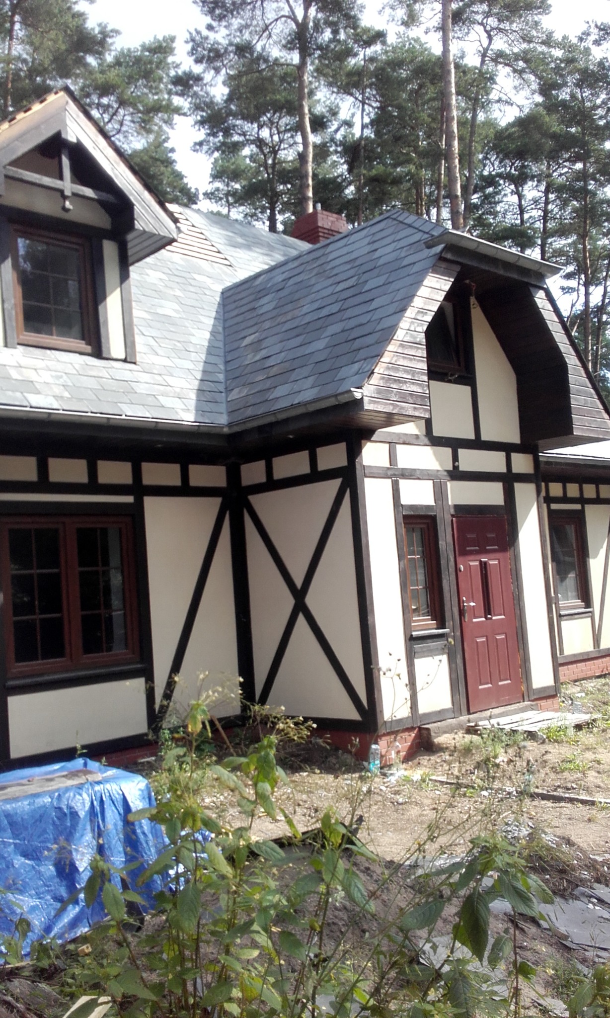 Dom w stylu szachulcowym z dwuspadowym dachem pokrytym szarym łupkiem, widok z poziomu gruntu, na pierwszym planie zarośla i fragment niebieskiej plandeki.