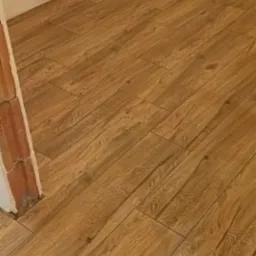 Ułożona podłoga z płytek ceramicznych imitujących drewno, widoczny fragment ściany i surowy mur przy futrynie drzwiowej.