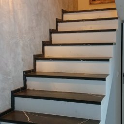 K. R. Firma remontowo - budowlana Radosław Kuchta - Schody wewnętrzne z czarnymi stopniami imitującymi marmur, białe podstopnice, ściana wykończona w stylu betonu architektonicznego, oświetlenie punktowe i obraz z motywem roślinnym w tle.