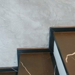 K. R. Firma remontowo - budowlana Radosław Kuchta - Schody z brązowego marmuru z białymi żyłami, przylegające do ściany z szarą dekoracyjną fakturą.