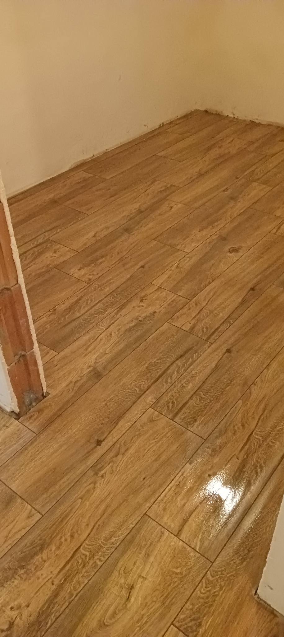 Ułożona podłoga z płytek ceramicznych imitujących drewno, widoczny fragment ściany i surowy mur przy futrynie drzwiowej.