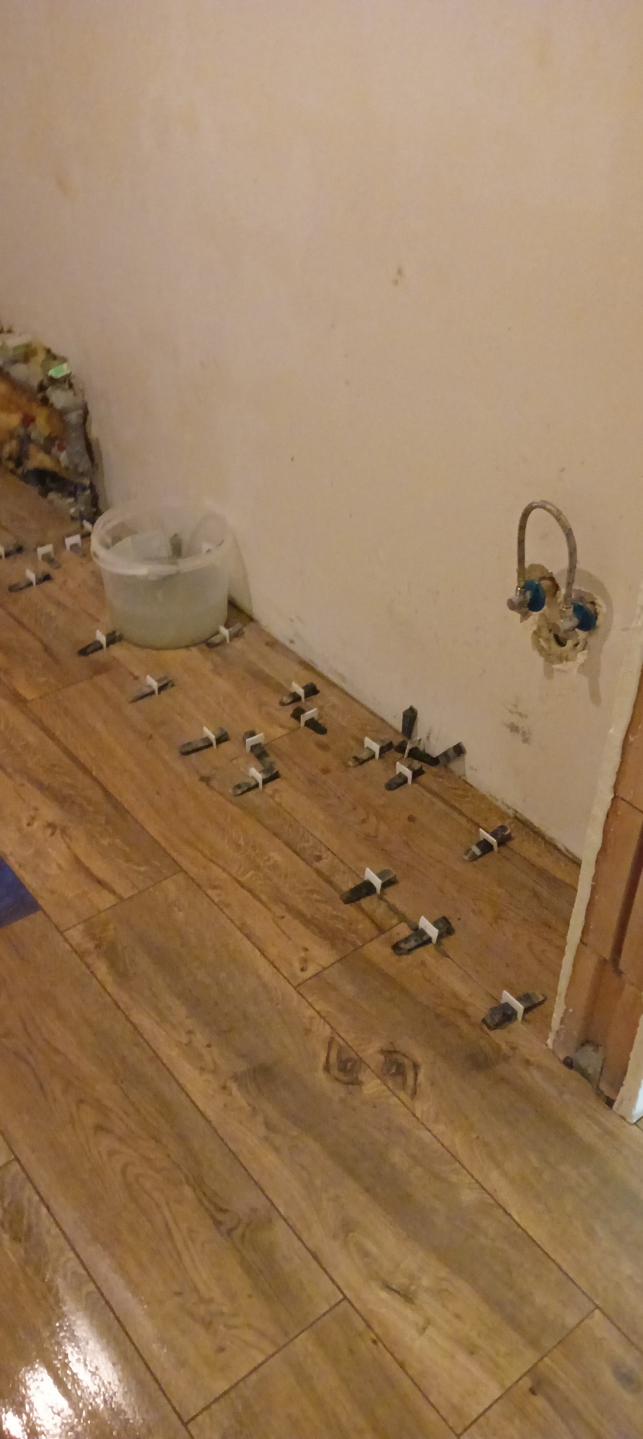 Układanie paneli podłogowych z użyciem krzyżyków dystansowych w rogu pomieszczenia; widoczna instalacja wodna na ścianie i wiadro z klejem.