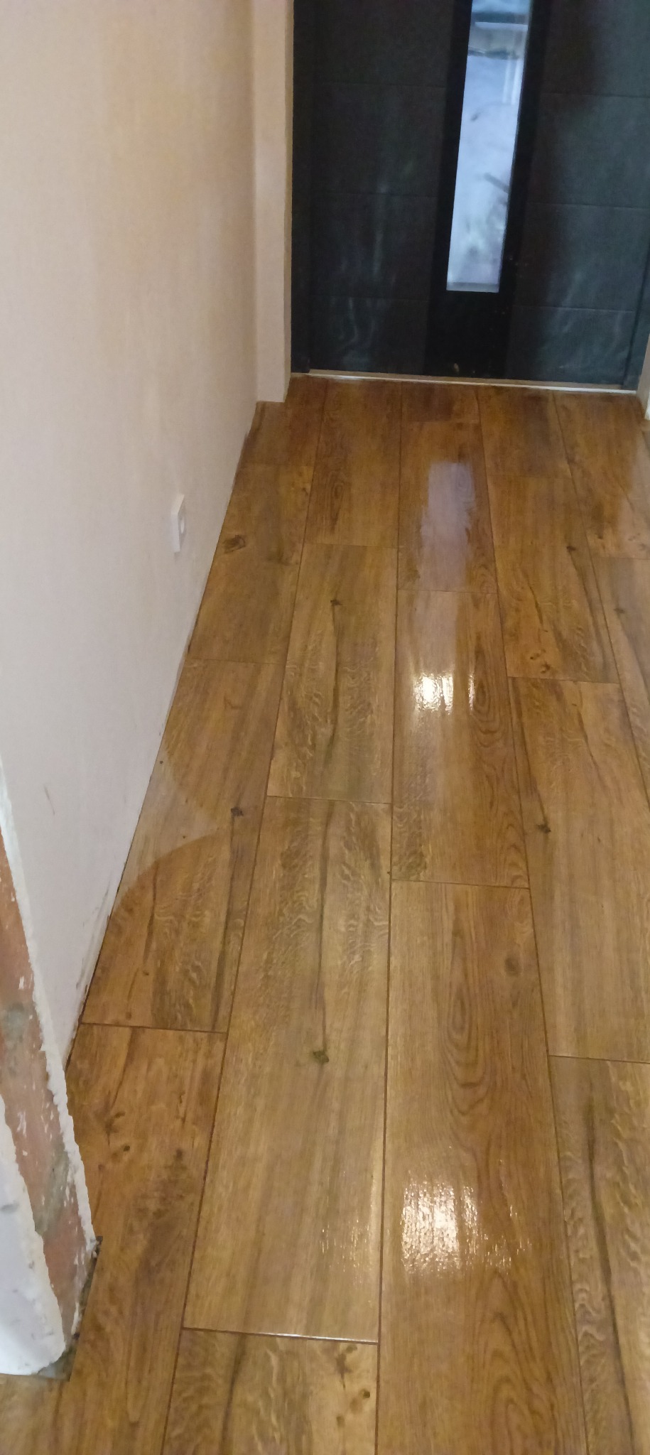 Ułożona, błyszcząca podłoga z paneli imitujących drewno, widoczny fragment ściany z gładzią i czarne drzwi.