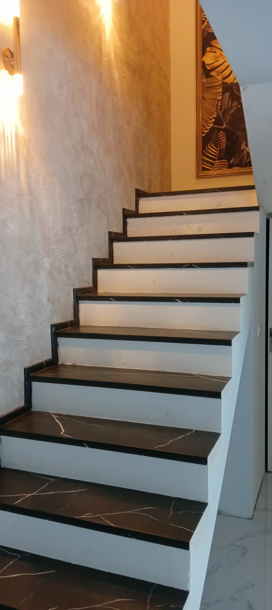 Schody wewnętrzne z czarnymi stopniami imitującymi marmur, białe podstopnice, ściana wykończona w stylu betonu architektonicznego, oświetlenie punktowe i obraz z motywem roślinnym w tle.