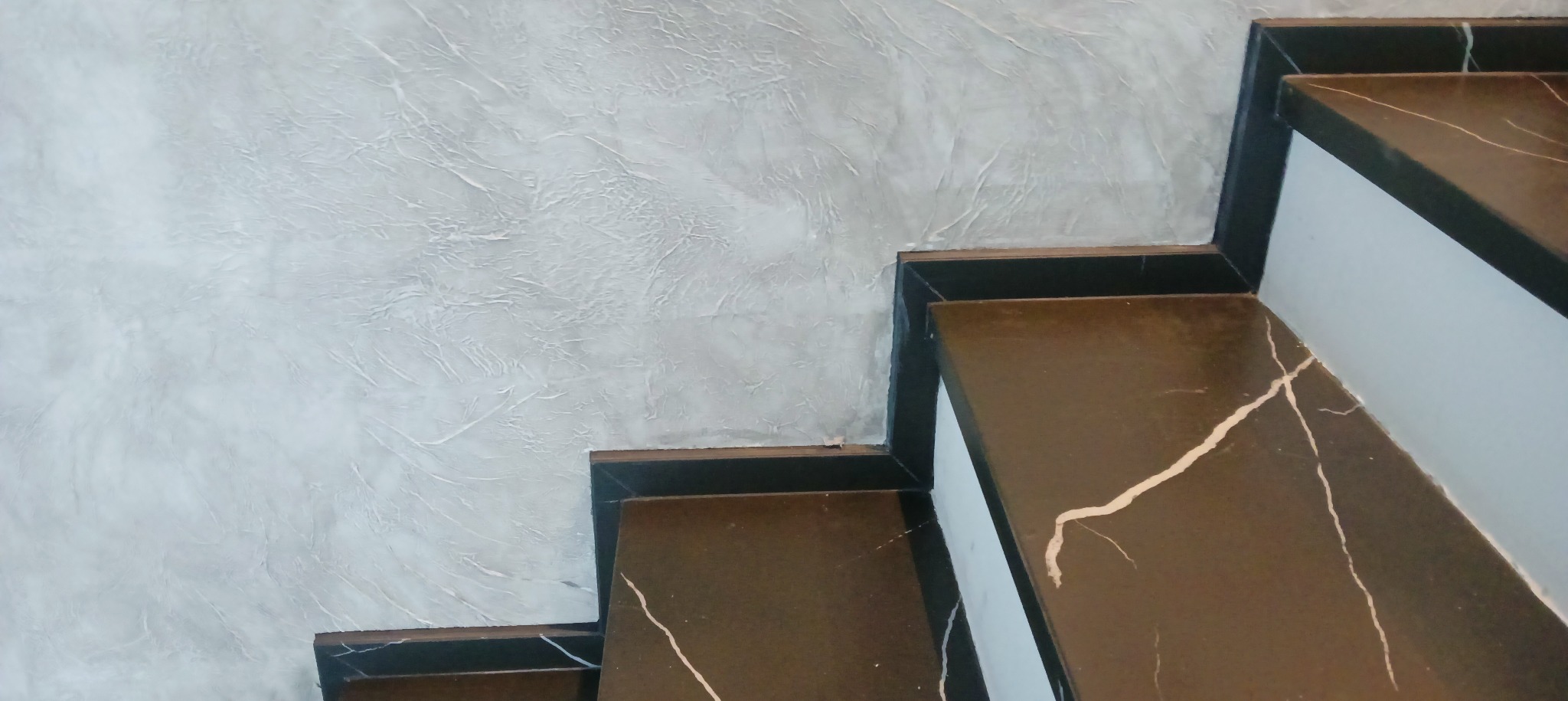 Schody z brązowego marmuru z białymi żyłami, przylegające do ściany z szarą dekoracyjną fakturą.