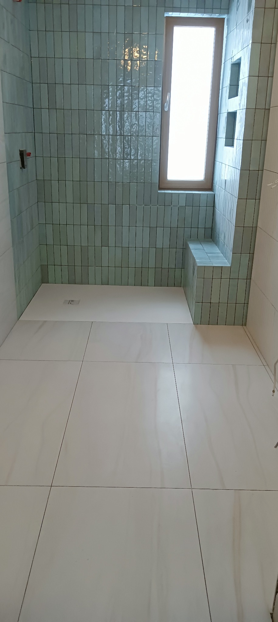 Świeżo wykafelkowana łazienka z turkusowymi, pionowo ułożonymi płytkami na ścianach i jasnymi, kwadratowymi płytkami na podłodze; widoczne okno z drewnianą ramą i wnęka wyłożona płytkami.