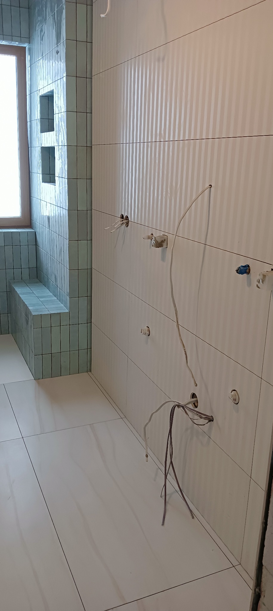 Łazienka w trakcie wykańczania: białe płytki z delikatnym wzorem pionowych pasków na ścianie, odkryte kable elektryczne, wnęki wyłożone turkusowymi płytkami, okno w tle.