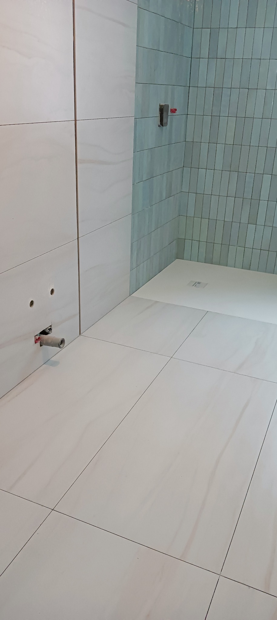 Wnętrze łazienki w trakcie prac wykończeniowych: białe płytki podłogowe i ścienne imitujące marmur, fragment ściany z niebieskimi, pionowo ułożonymi płytkami, widoczne instalacje...