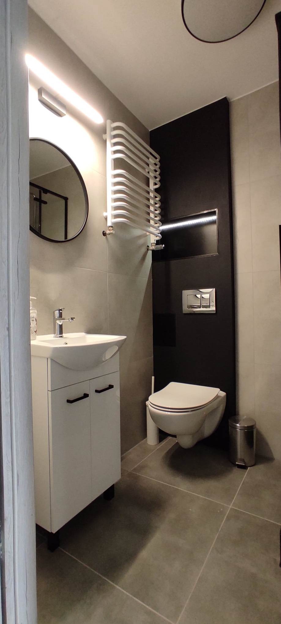 Nowoczesna łazienka z szarą płytką, białą umywalką z szafką, podwieszaną toaletą na czarnym tle i grzejnikiem drabinkowym.